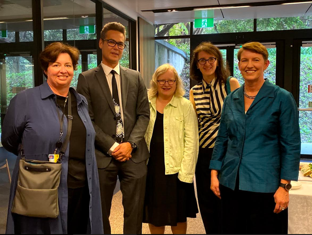 Lisa McIntosh, Peter Chen, Pip Pattison, Agata Mrva-Montoya, Susan Murray standing from left to right smiling at the camera in the basement of University of Sydney's Fisher Library. A glass door is in the background with leafy surroundings.