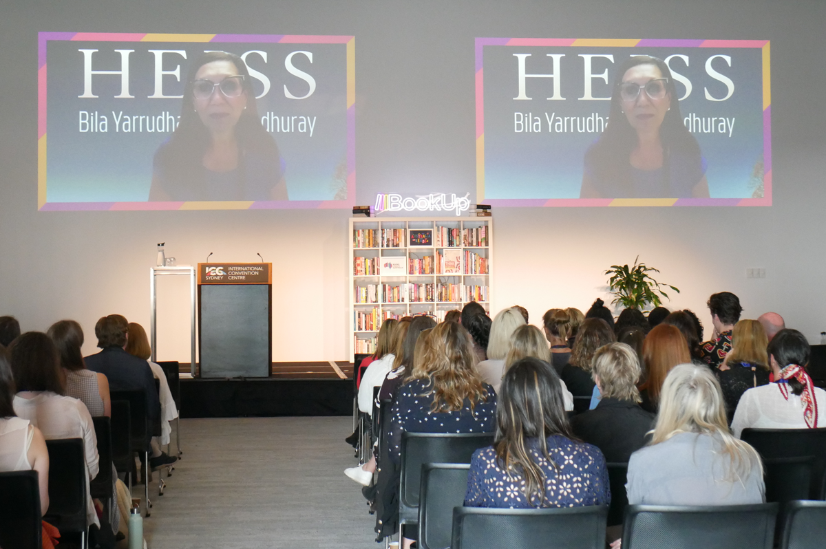 A photograph of Anita Heiss delivering the opening address for the 2021 BookUp conference via video link. She is projected onto two screens above a stage with an empty podium. In the front of the frame, the physical BookUp audience sits in rows of chairs.