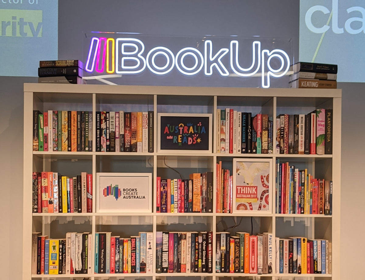 A photograph: A neon sign of the BookUp logo stands between piles of books atop a white bookcase. The logo is 'BookUp' in white sans serif font and three vertical lines in purple, pink, and orange, stylised as books leaning against the 'B'. The bookcase shelves are packed with Australian books, as well as three posters, for Australia Reads, Books Create Australia, and Think Australian 2017.