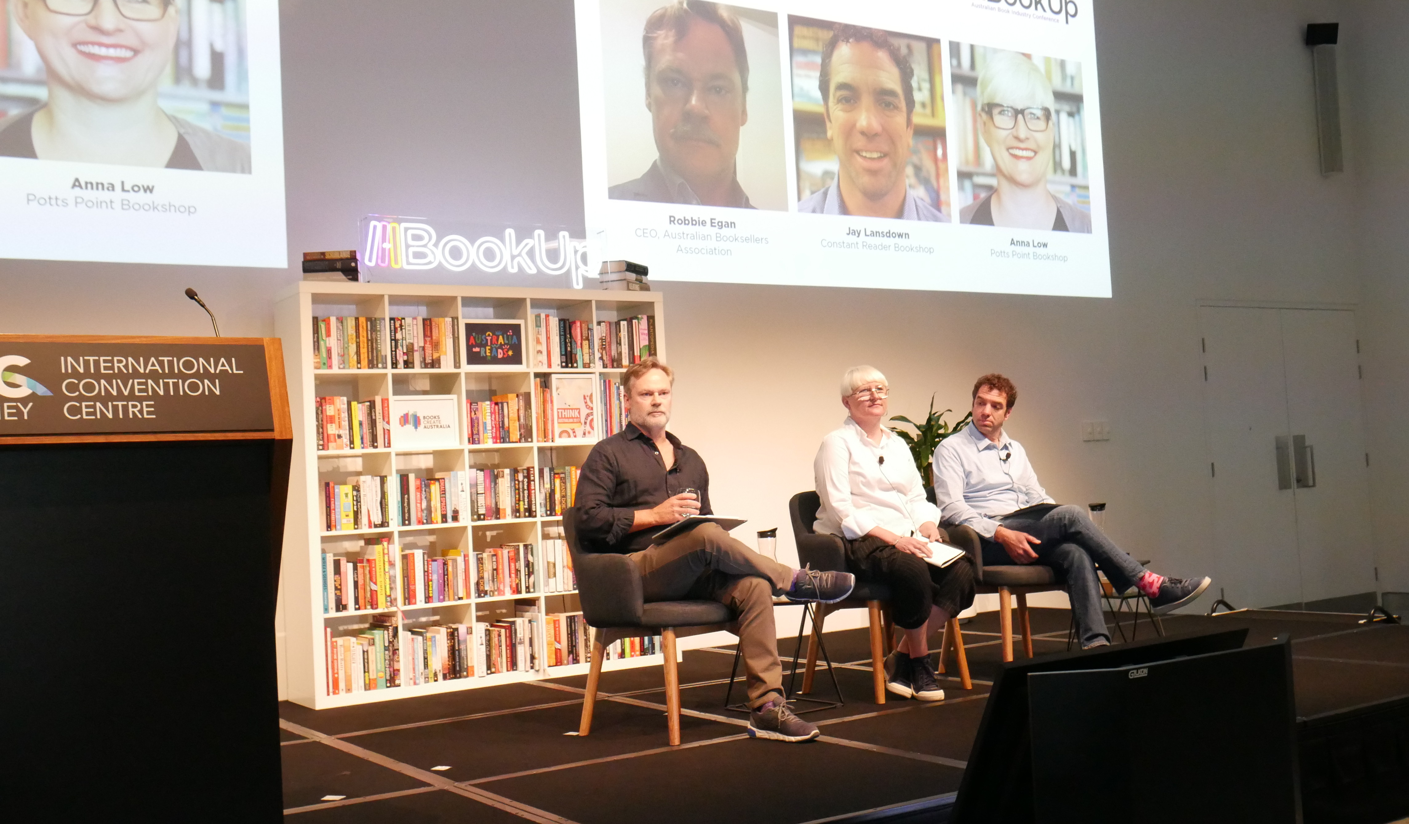 A photograph of two men and a woman, sitting in chairs on the BookUp stage. Projected onto the wall behind them is a slide which reads 'The Future for Bookshops' above headshots of the speakers.