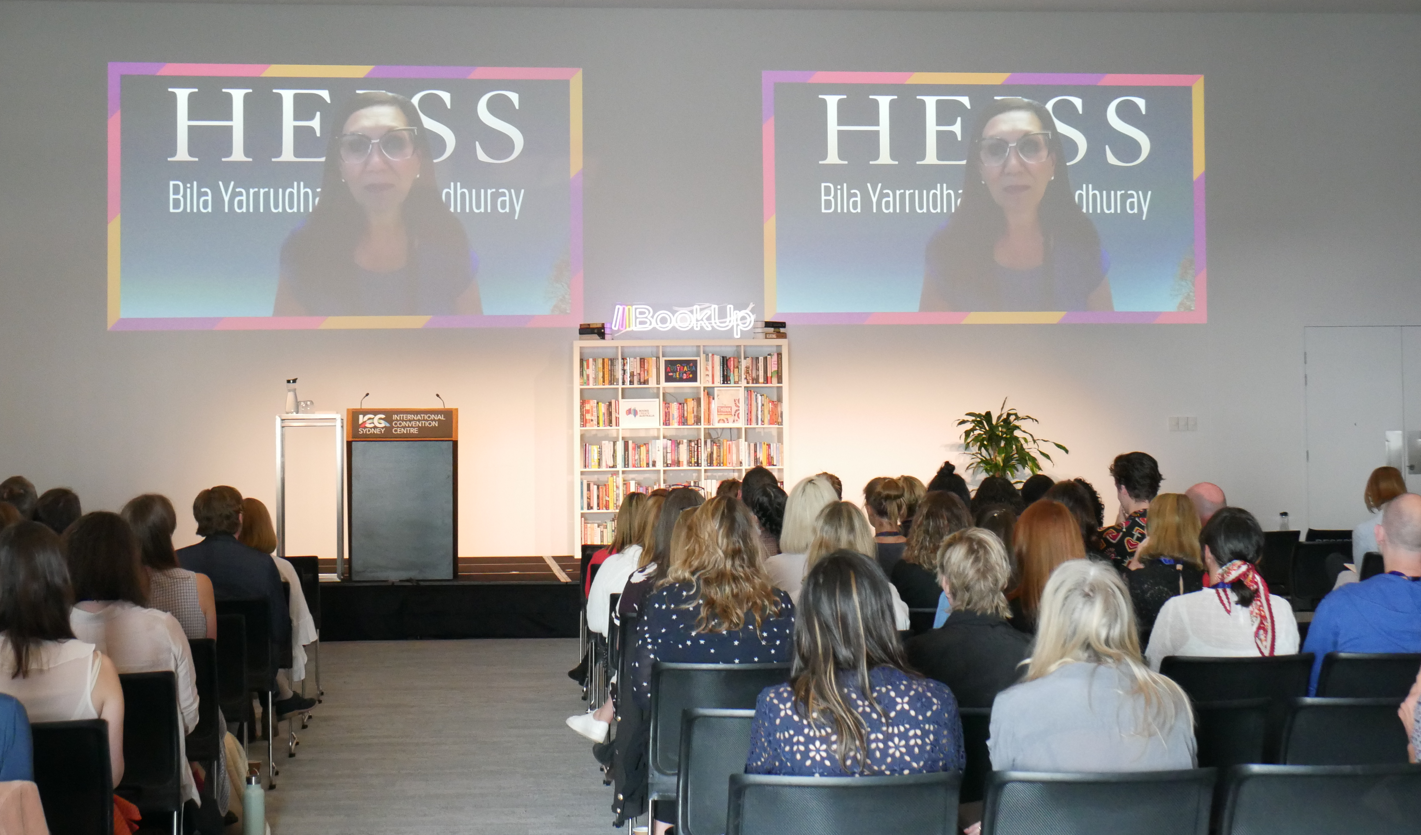 A photograph of Anita Heiss delivering the opening address for the 2021 BookUp conference via video link. She is projected onto two screens above a stage with an empty podium. In the front of the frame, the physical BookUp audience sits in rows of chairs.