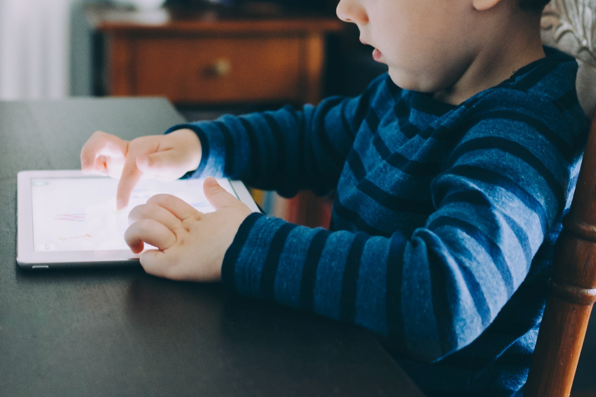 A photograph of a child using both hands to interact with a tablet at a table.